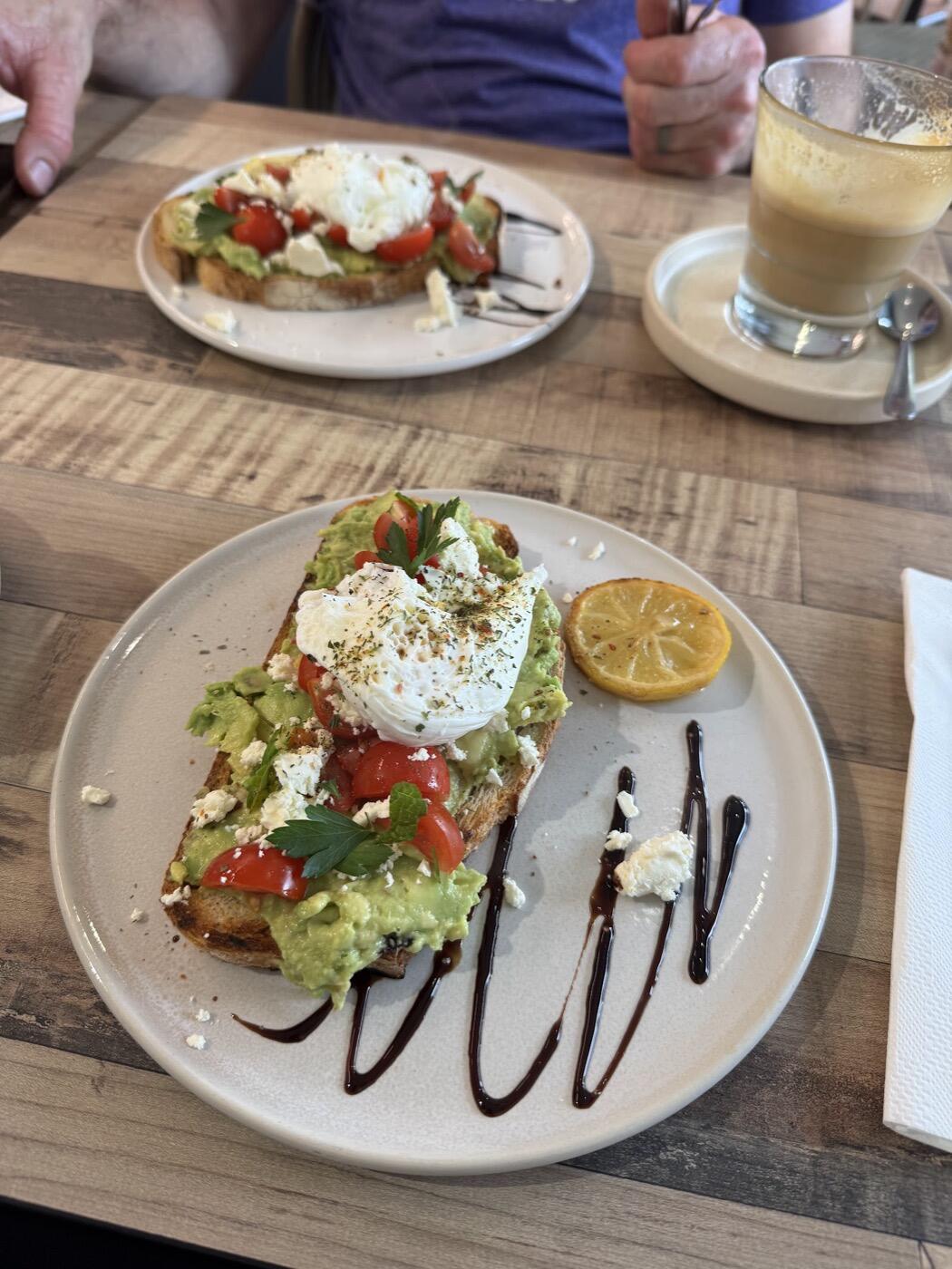Smashed avocado on toast topped with a poached egg, tomato and feta, with a lemon wedge, on a round plate drizzled with balsamic
