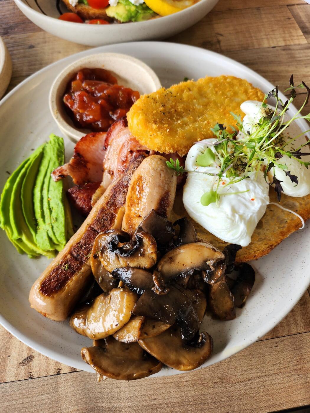 A big breakfast plate with a sausage, bacon, garlic mushrooms, a hash brown, avocado, a poached egg with micro herbs and a pot of relish