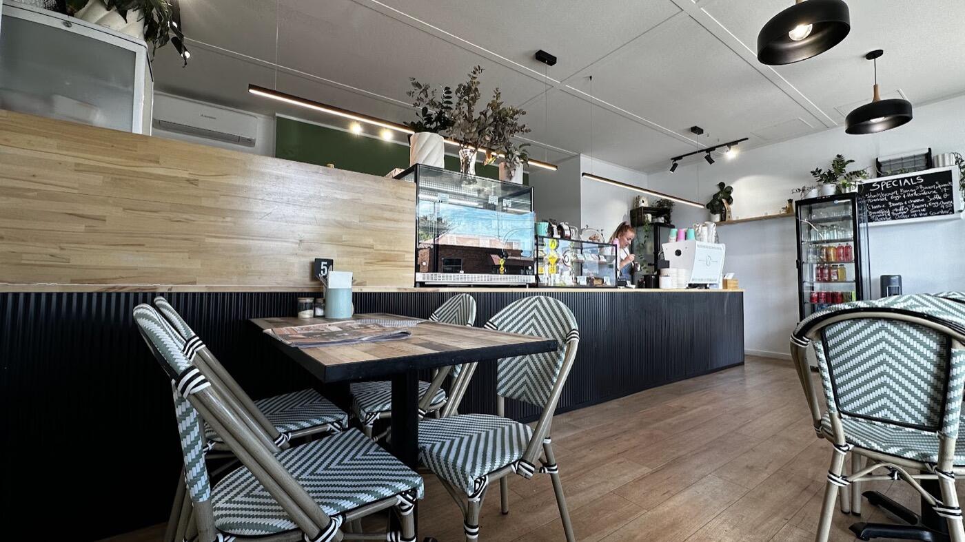 Inside the café: bistro chairs with black and white chevron seats at a timber table, a dark counter with pendant lights above and a timber floor