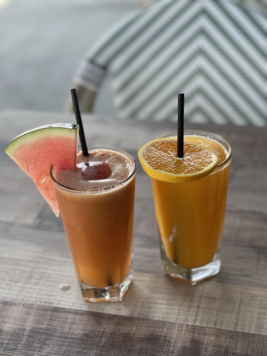 Two tall glasses with straws, one orange and one amber, with a wedge of watermelon on the rim of one