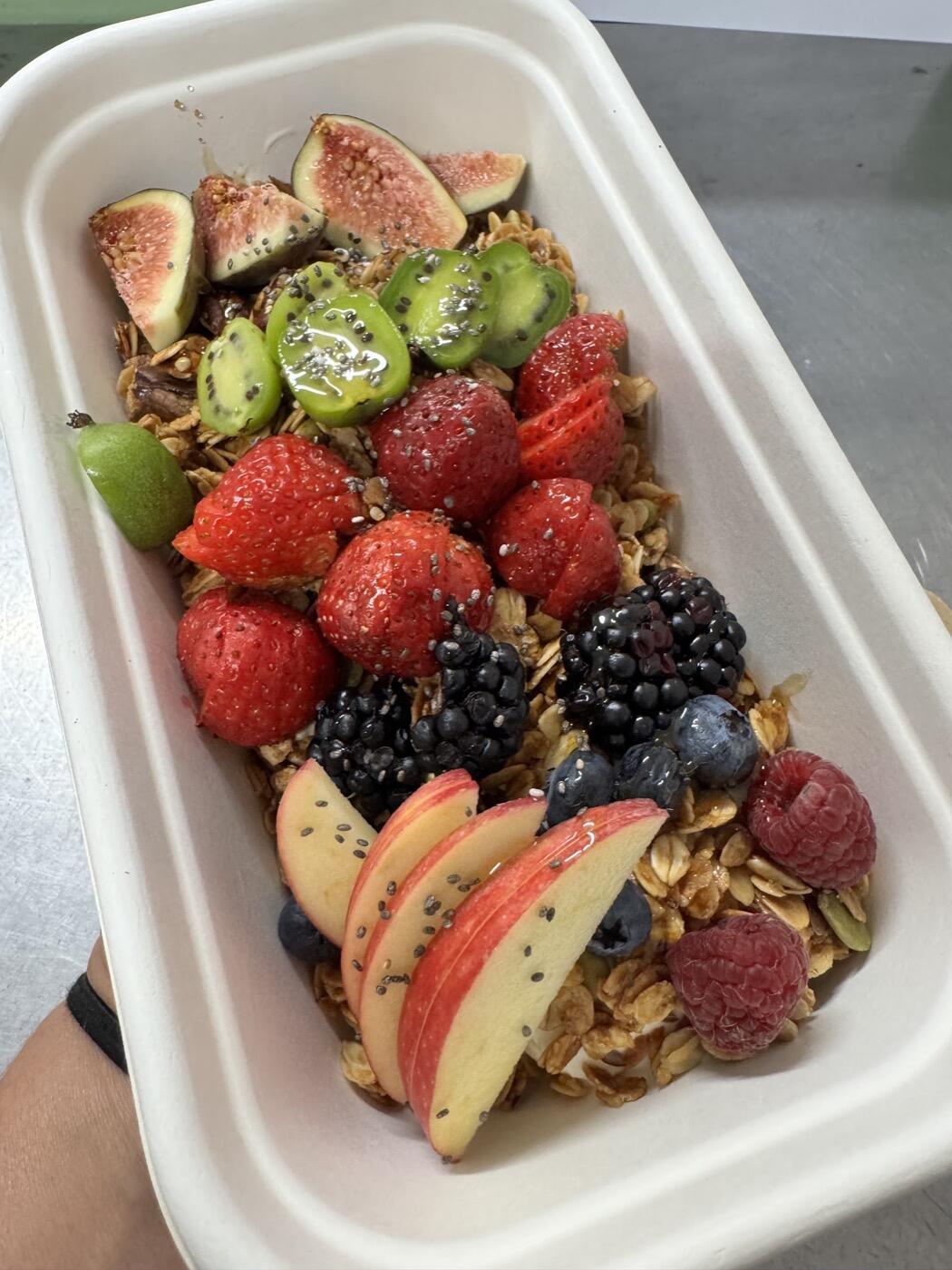 A takeaway box of granola topped with strawberries, blackberries, blueberries, kiwi fruit, fig and apple slices