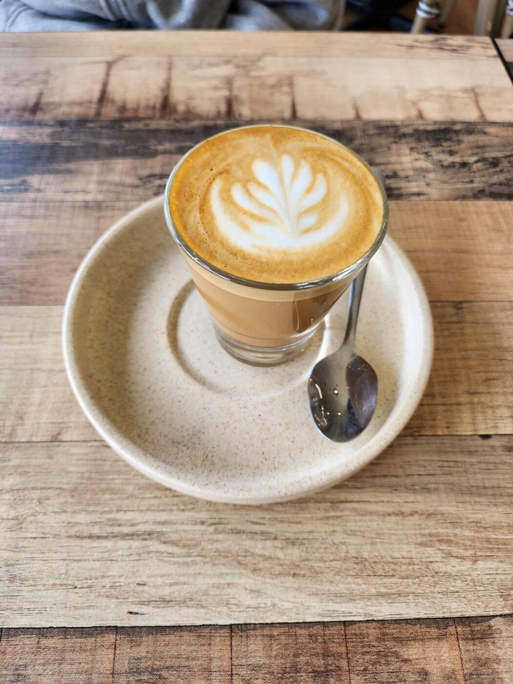 A latte with a rosetta pattern in a glass cup on a saucer, on a timber table
