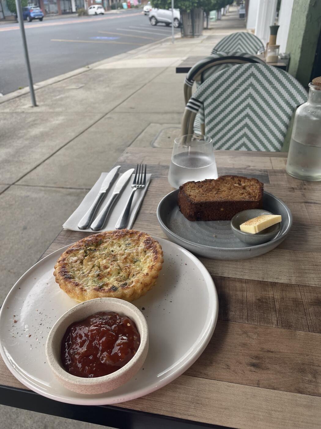 A savoury quiche beside a pot of red relish on a plate, with a slice of loaf and a pat of butter on a plate behind