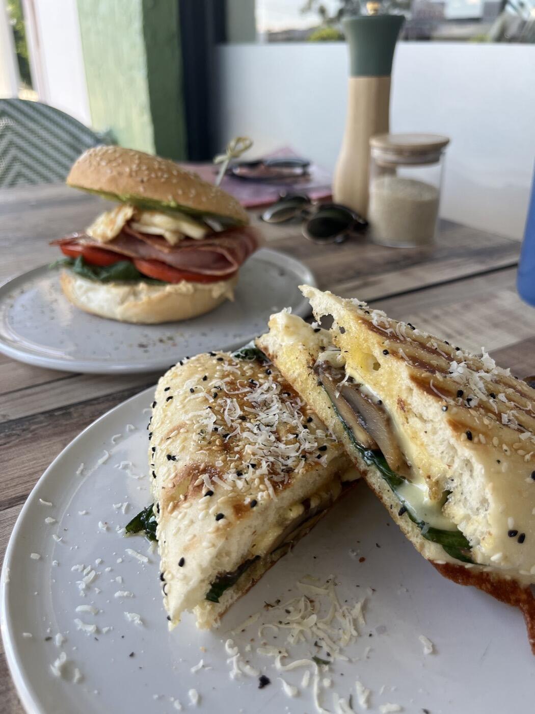 A toasted bagel sandwich cut in half with melted cheese and greens, and a filled roll on a plate behind it